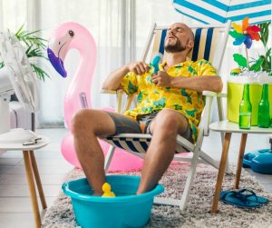 Guy wearing a Hawaiian shirt and shorts in a beach chair looking too hot with his feet in a small tub, cold beverages and fans blowing on him.