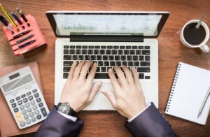Business professional working on a laptop at a desk with calculator, notebook, pens, and coffee, representing creative ways to manage finances when money is tight.